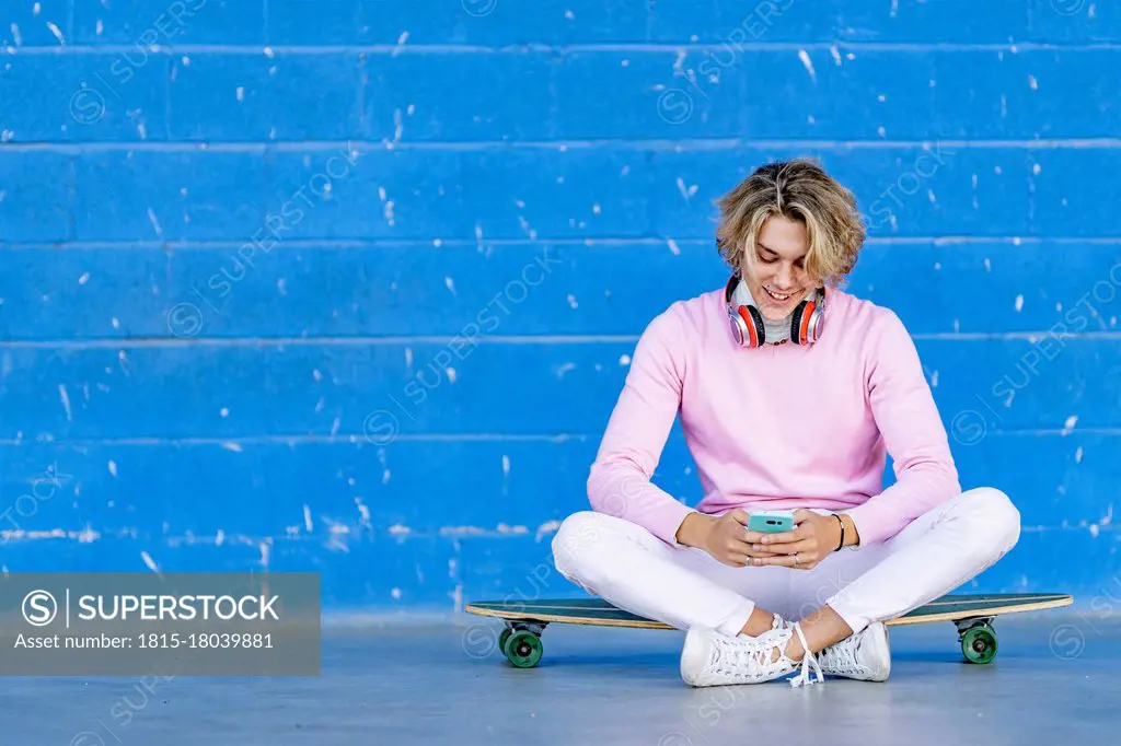 Blond man using phone while sitting on skate board against blue wall