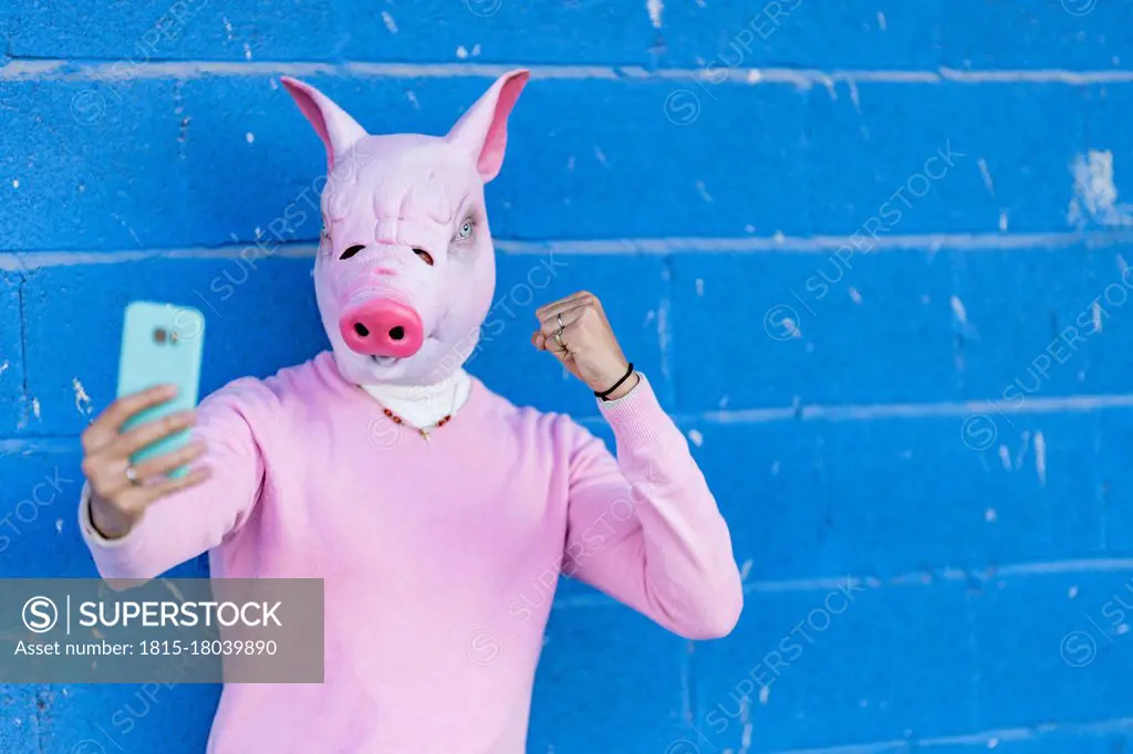 Young man in pig mask showing fist while taking selfie against blue wall