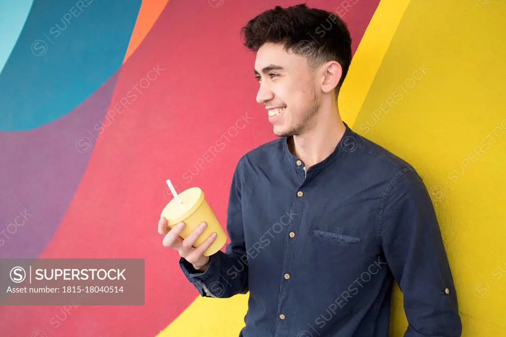 Happy young man holding disposable cup while looking away against multi colored wall