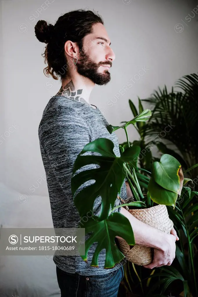 Young man looking away while holding Monstera plant at home