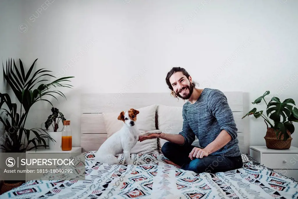 Handsome man playing with dog while sitting on bed at home