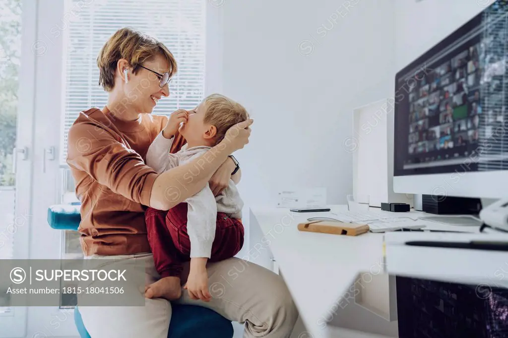 Loving mother embracing son sitting on her lap at home office