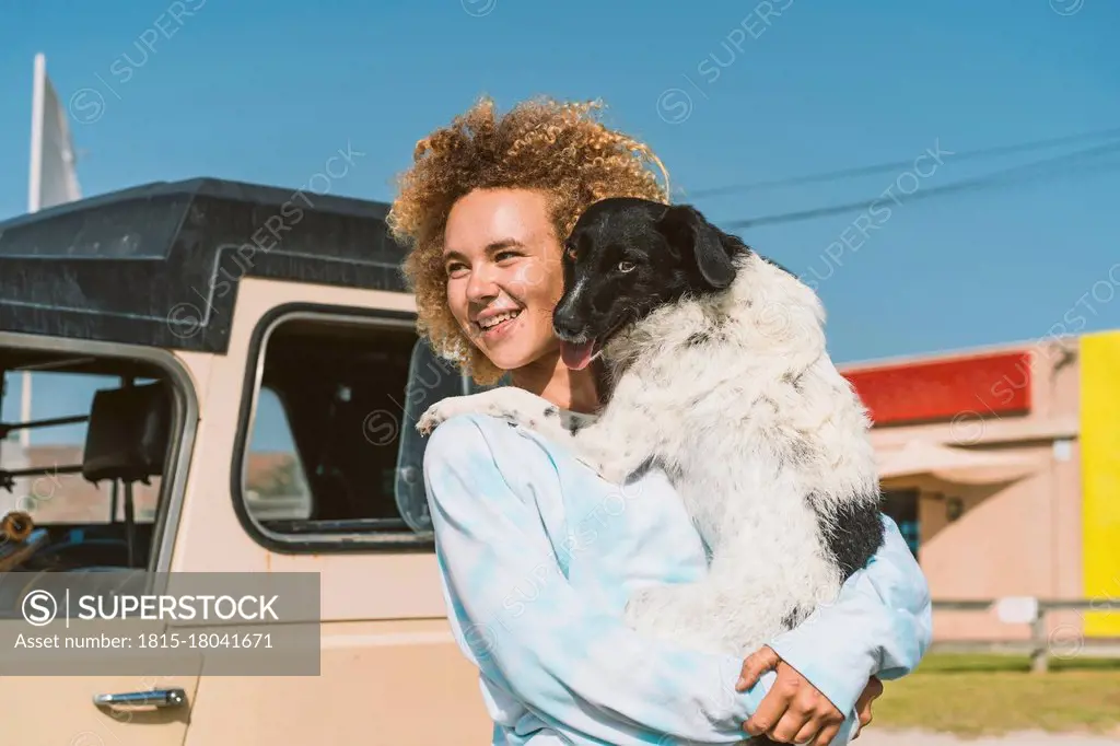 Smiling young blond woman carrying dog while looking away on sunny day