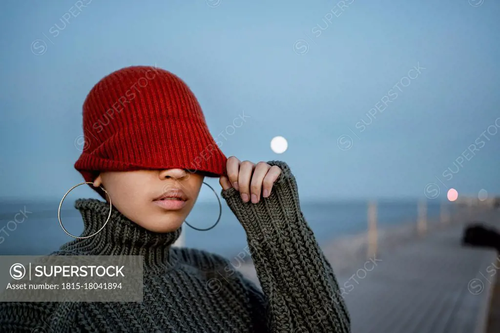 Woman covering eyes with knit hat while standing against sky