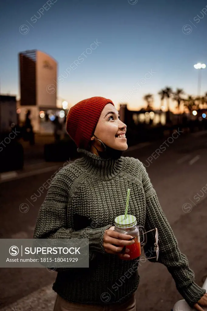 Smiling woman with face mask below chin holding juice while standing on road