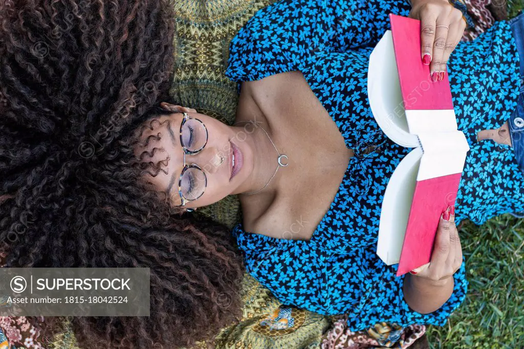 Afro young woman with curly hair reading book while lying down at public park