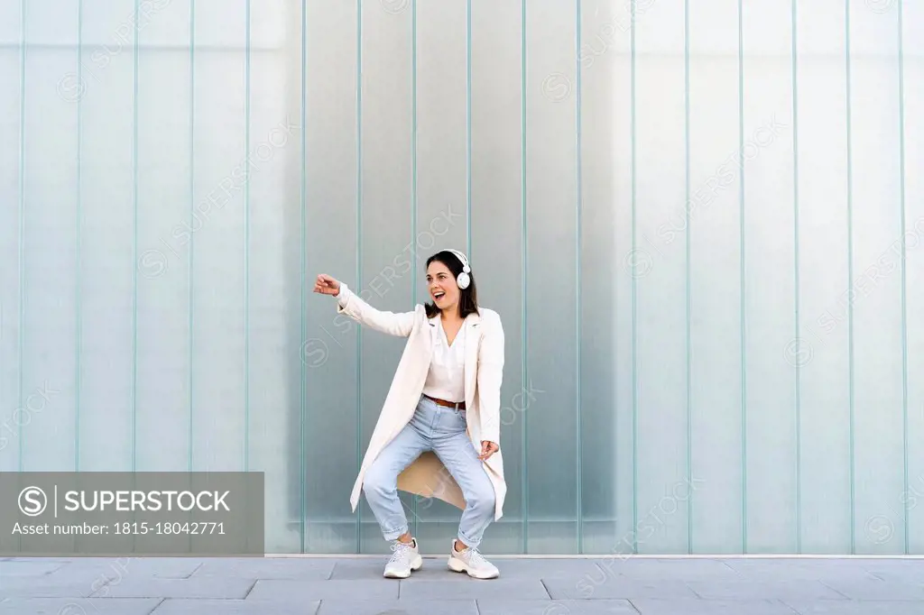 Cheerful businesswoman listening music through headphones while dancing on footpath against glass wall