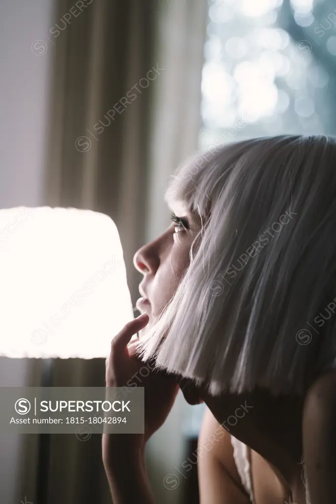 Young woman with hand on chin standing by illuminated lamp at home