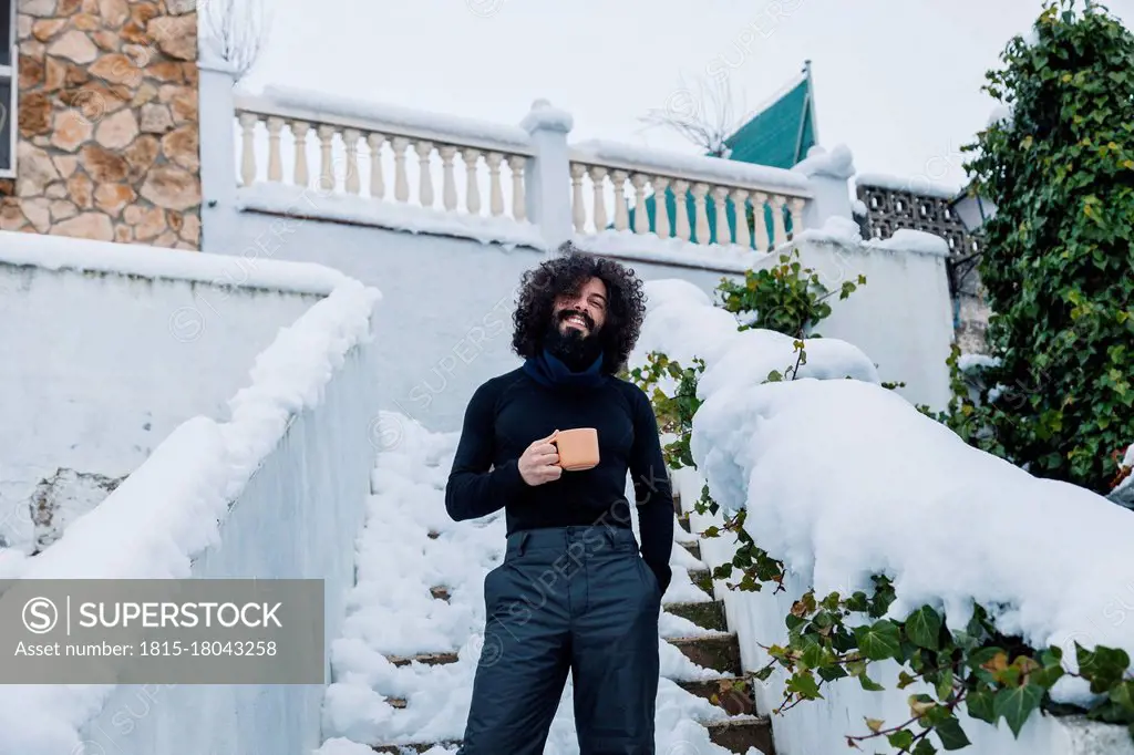 Smiling bearded man with coffee cup standing on snow covered steps