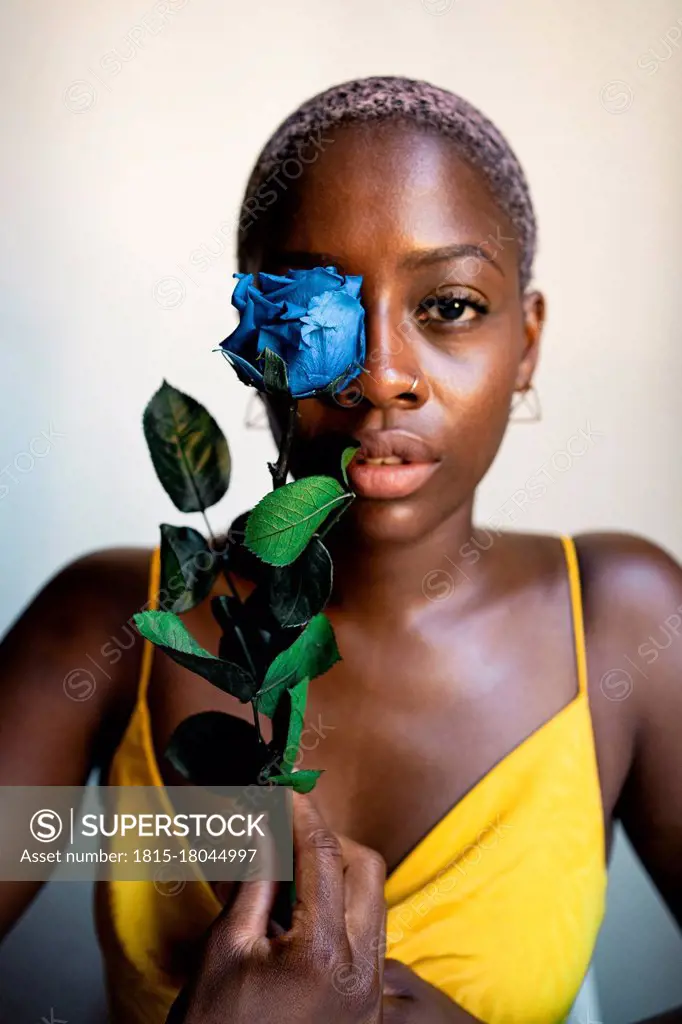 Confident woman holding blue rose in front of eye at home