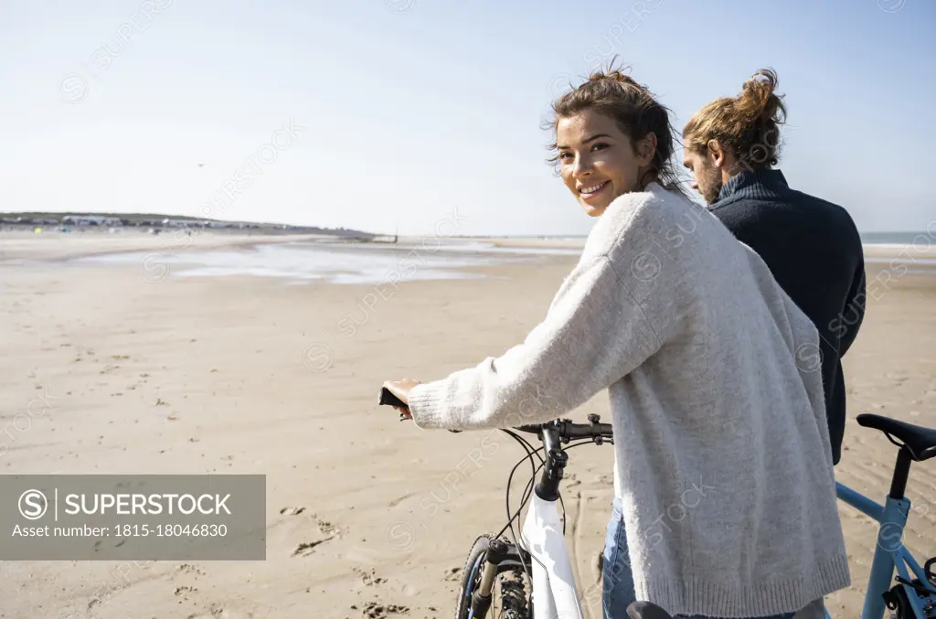 Smiling beautiful woman walking with bicycle by boyfriend while looking over shoulder at beach against clear sky on sunny day