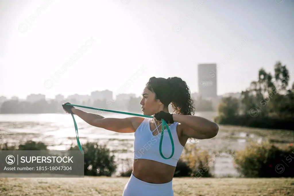 Female athlete stretching resistance band while exercising in public park against clear sky