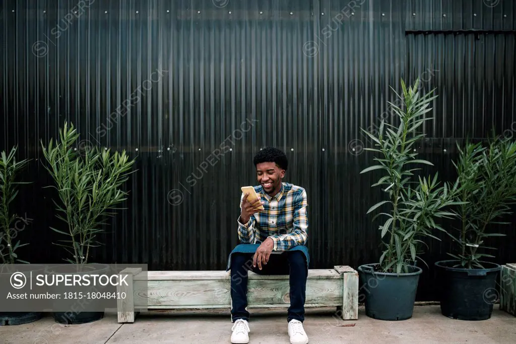 Smiling male worker using mobile phone while sitting on bench against corrugated wall