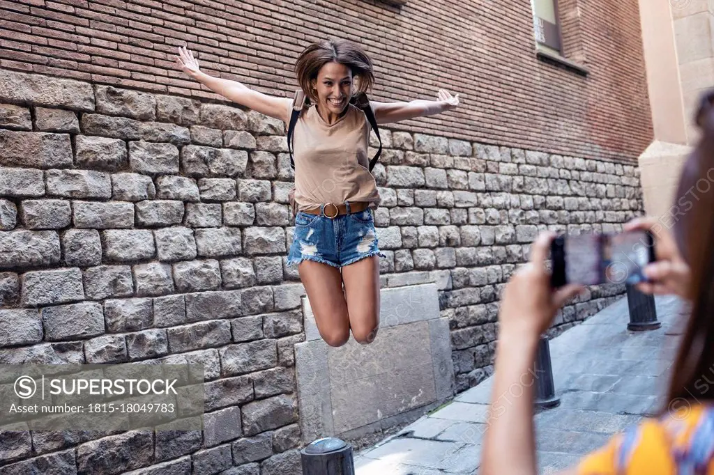 Friend taking photo of woman jumping against wall at Gothic Quarter in Barcelona, Catalonia, Spain