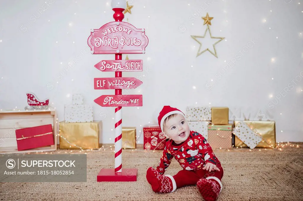 Smiling baby girl wearing Santa hat playing while sitting by direction pole at home during Christmas