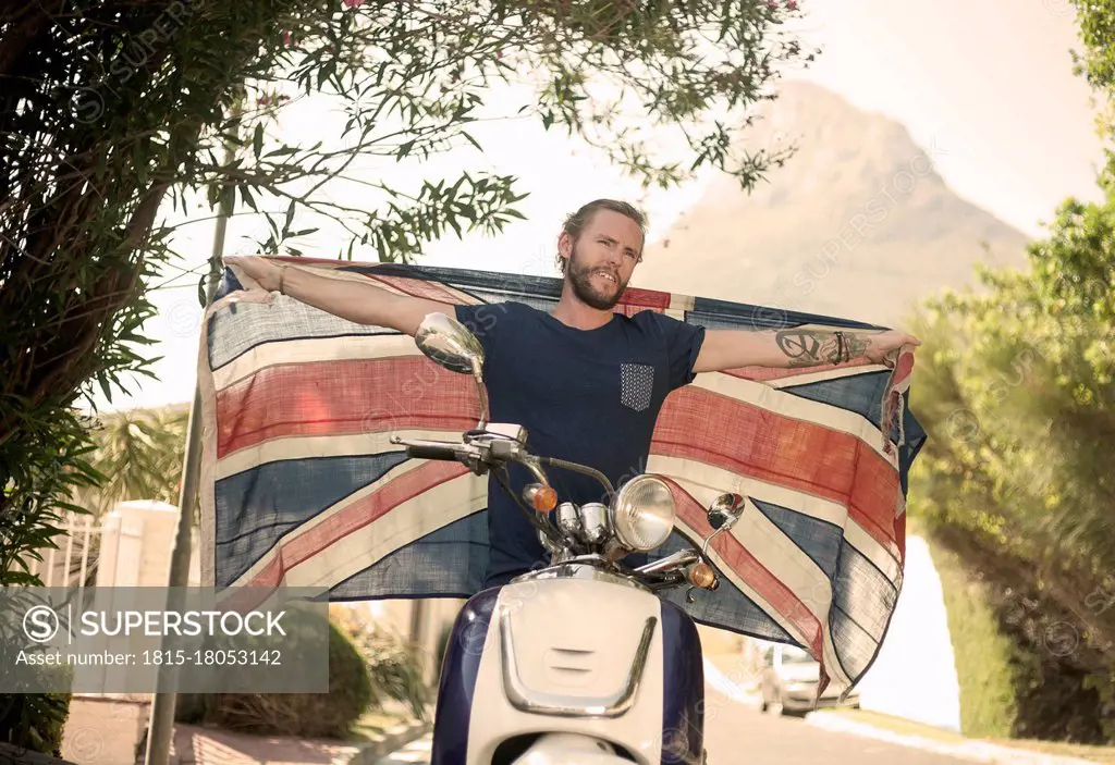 Man with arms outstretched holding national flag while sitting on motorcycle during summer