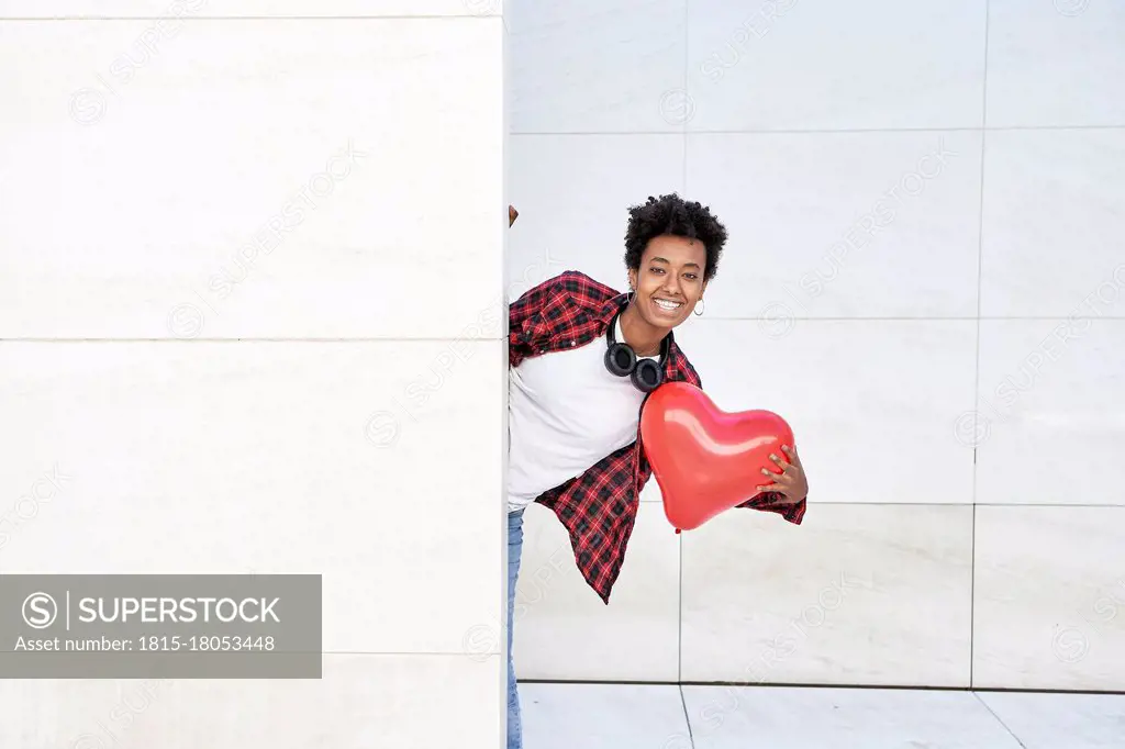 Happy young woman holding red heart shape balloon while standing behind white wall