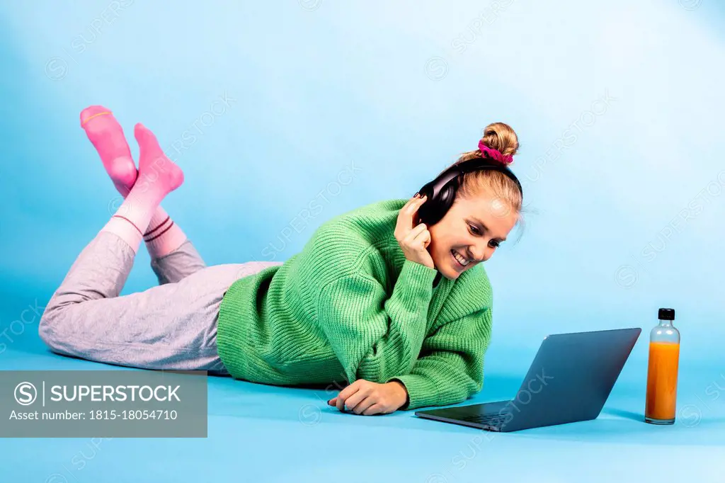 Smiling woman with headphones looking at laptop while lying on front against blue background