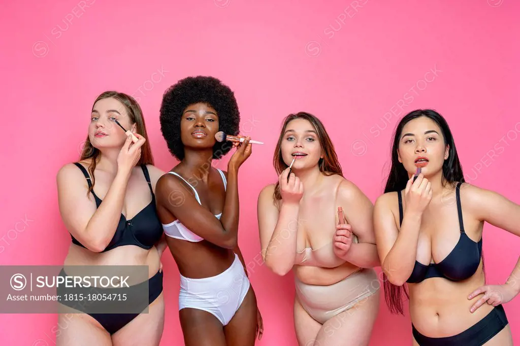 Multi-ethnic group of female models in lingerie applying make-up while standing against pink background