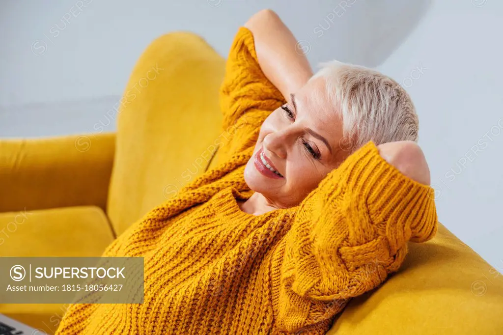 Smiling woman with hands behind back looking away while resting on sofa