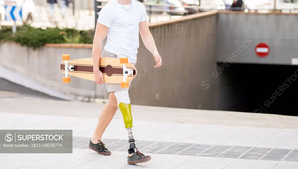 Young disabled man holding skateboard while walking on footpath