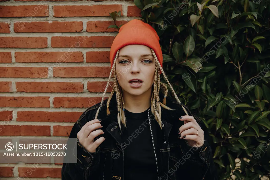 Beautiful woman wearing orange knit hat standing against brick wall and plants