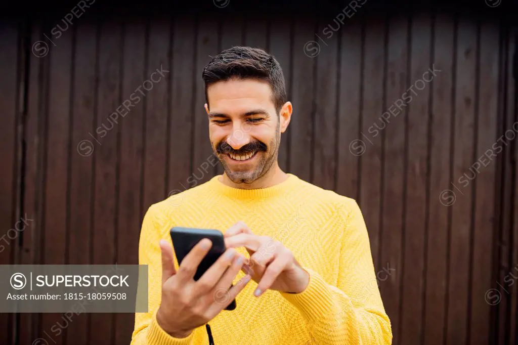 Smiling mid adult man using mobile phone against wooden wall