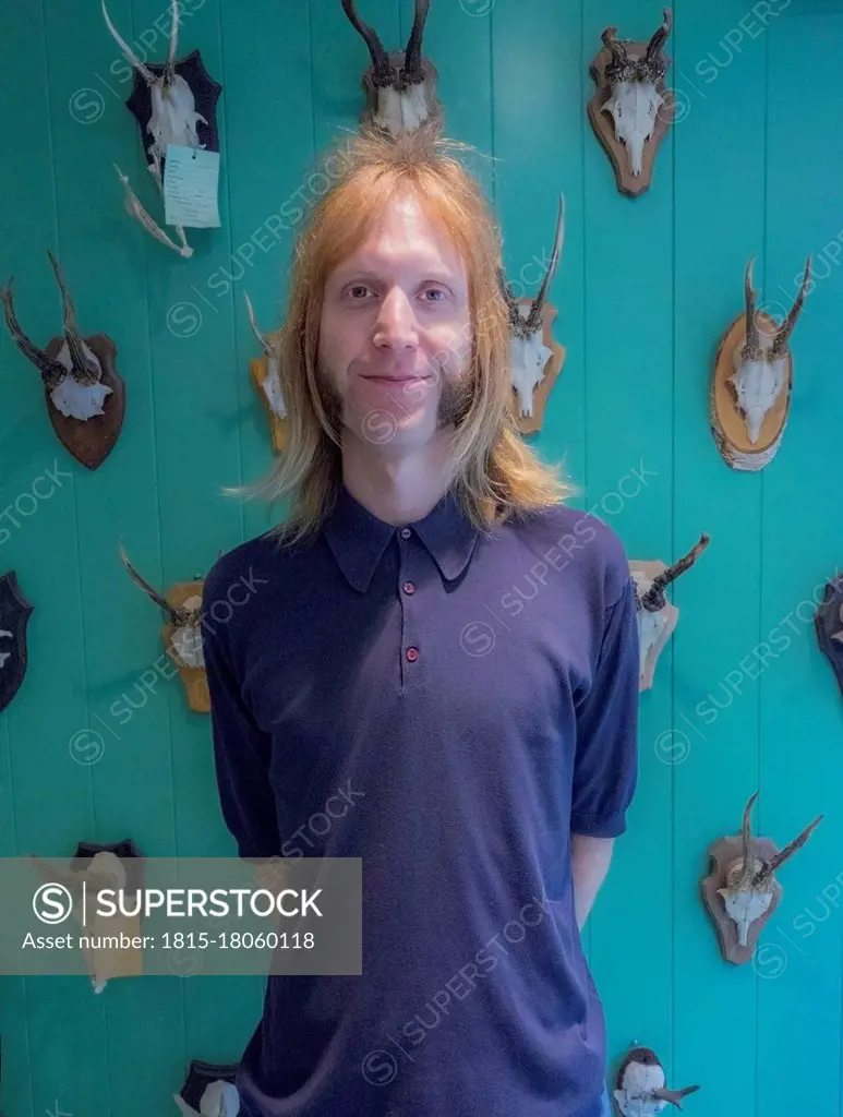 Blond male barber standing against animal skulls and antlers on turquoise wall