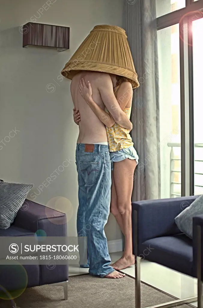 Young couple with lamp shade on head embracing each other while standing at home