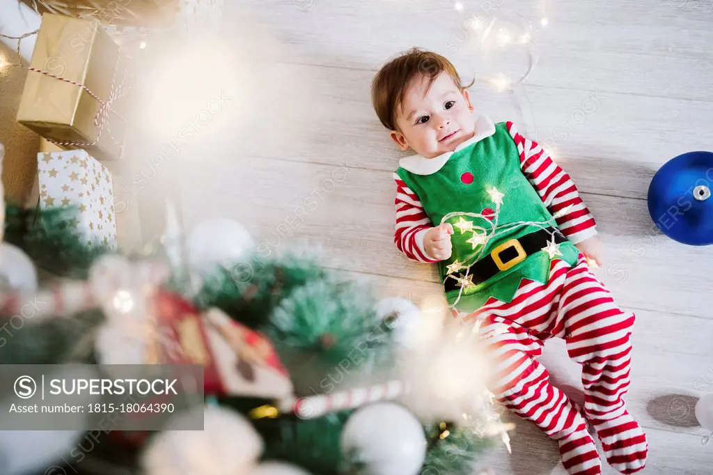 Baby boy in elf costume playing with string light while lying on floor at home during Christmas