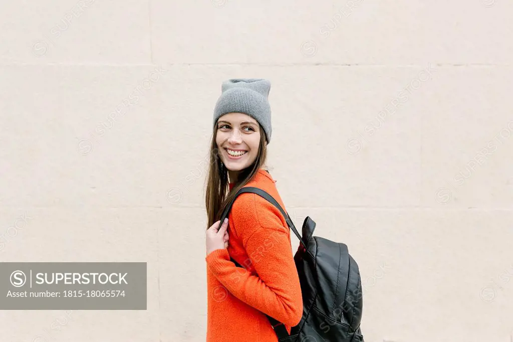 Smiling young woman with backpack looking away in front of wall