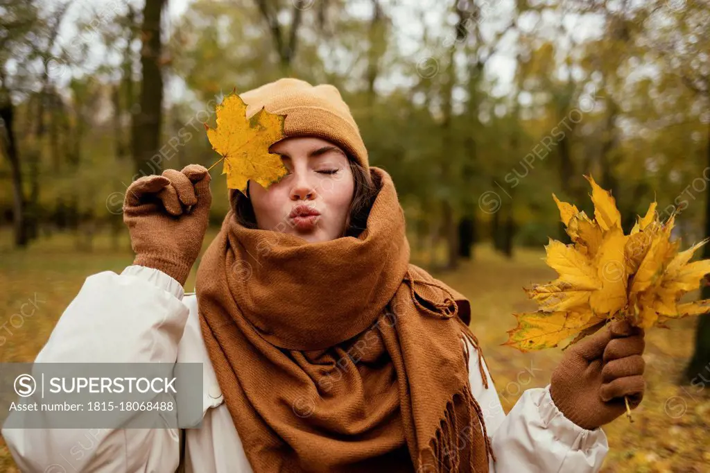 Young woman with eyes closed puckering while covering eye with autumn leaf in public park