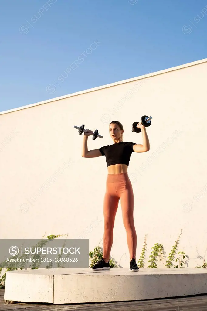 Sportswoman flexing muscle with dumbbell standing on pedestal against wall during sunrise