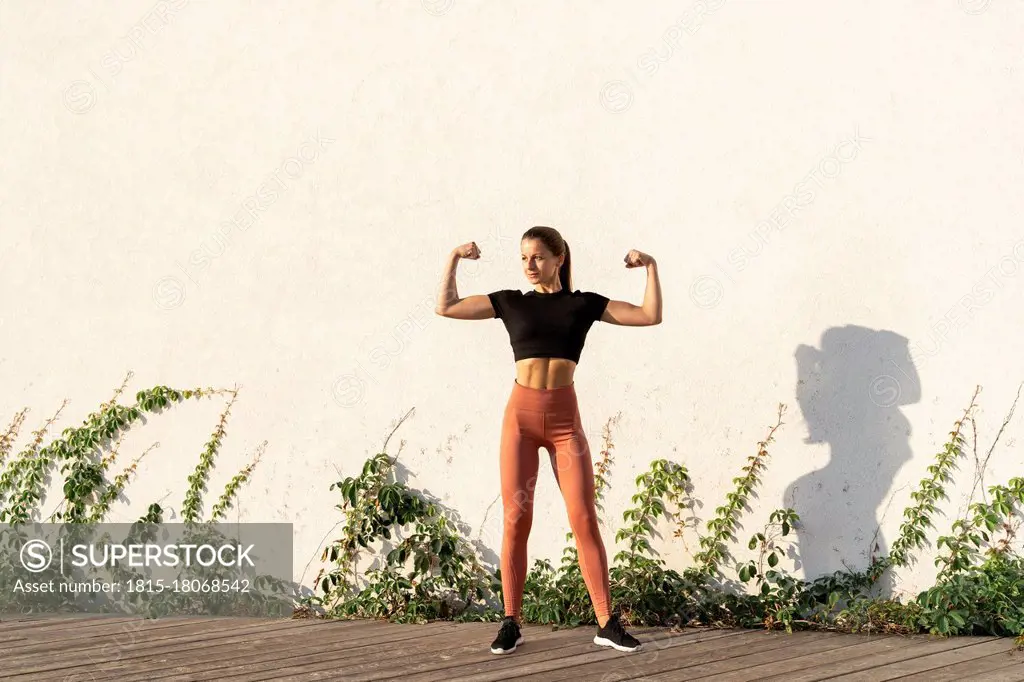 Young female athlete flexing muscle while standing against wall during sunrise