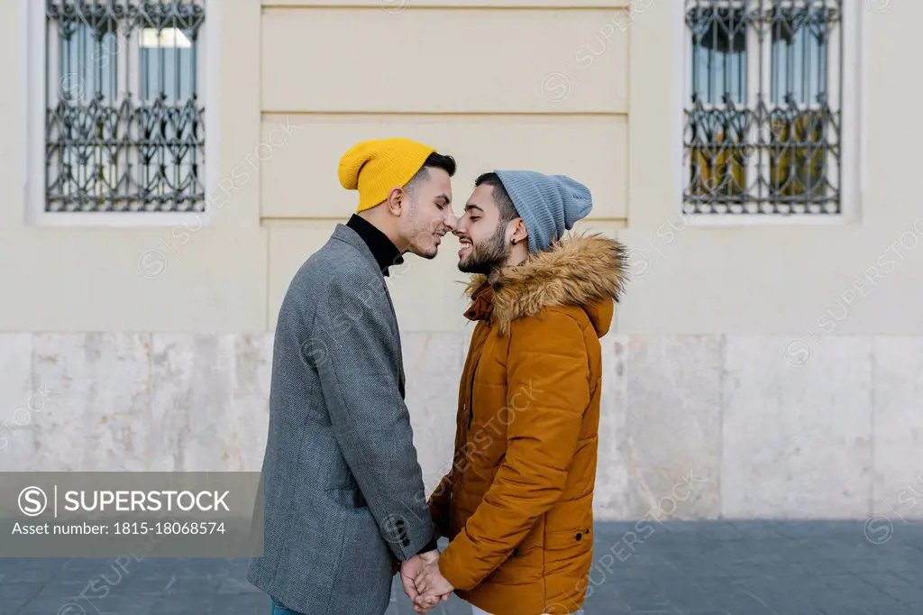 Gay men wearing jacket rubbing noses while holding hands standing against wall