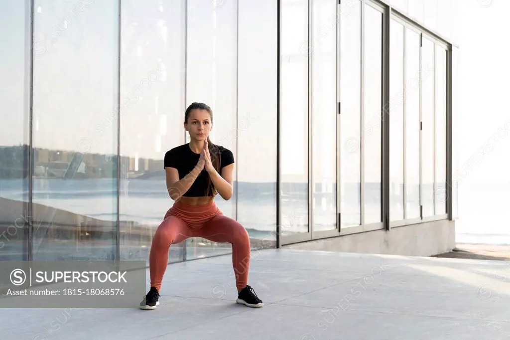 Young sportswoman doing squat exercise on promenade against glass window