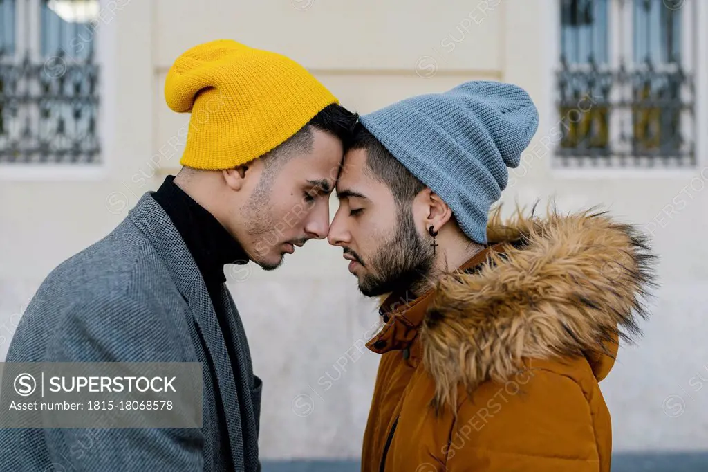 Gay couple wearing knit hat standing face to face with eyes closed against wall