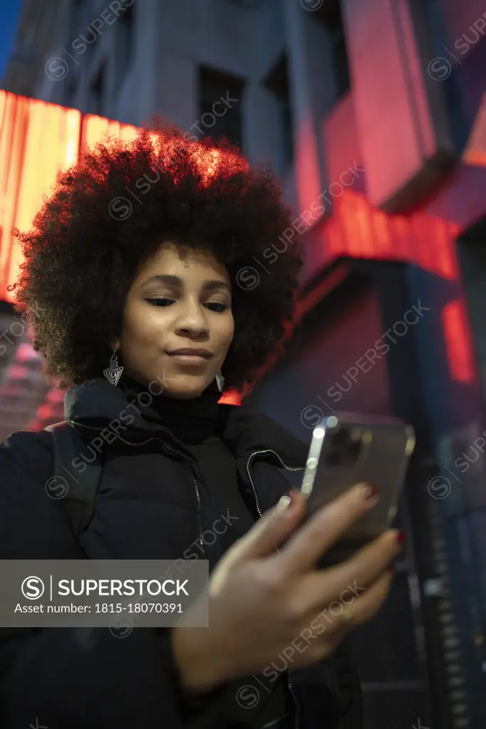 Afro young woman in warm clothing using smart phone in city at night