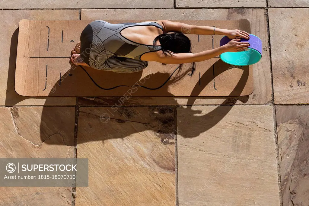 Female athlete practicing childs pose with yoga wheel on mat during sunny day