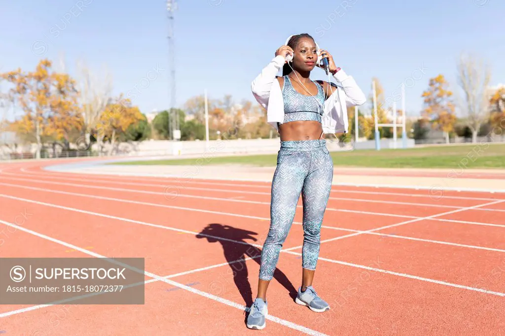 Female athlete wearing headphones while standing on sports track during sunny day