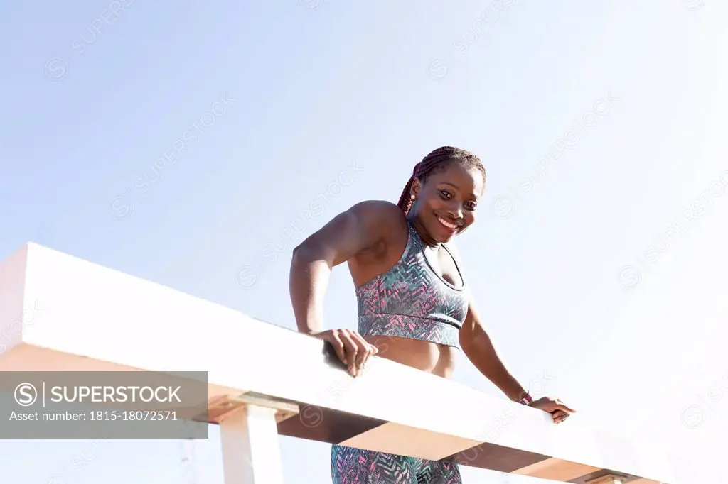 Happy sportswoman hanging on balance beam against clear sky during sunny day