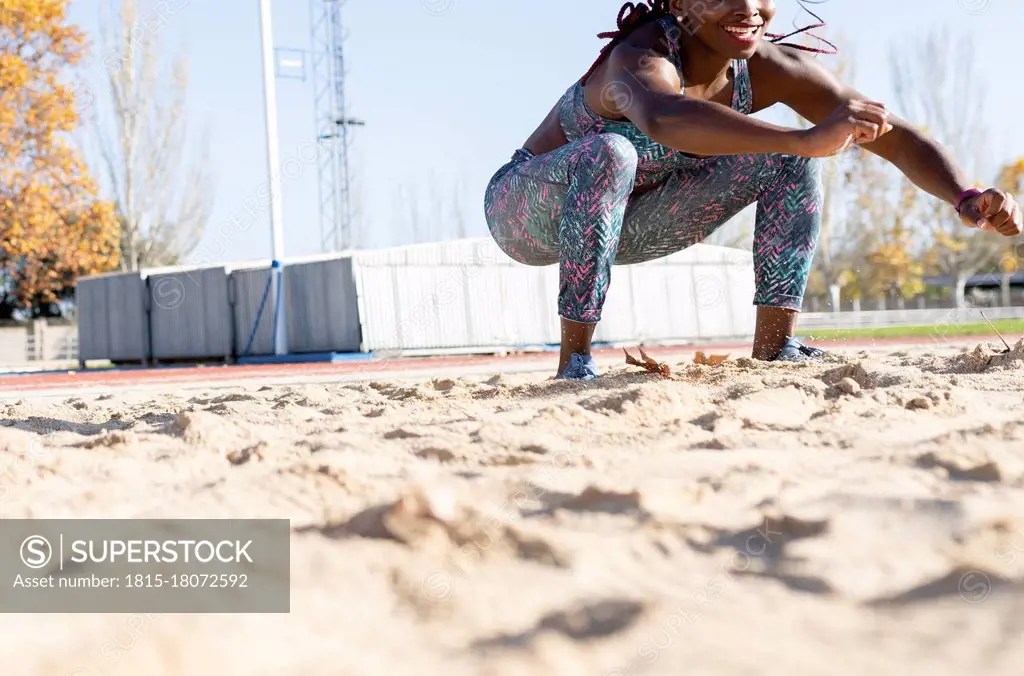 Smiling female athlete landing on sand in long jump during sunny day