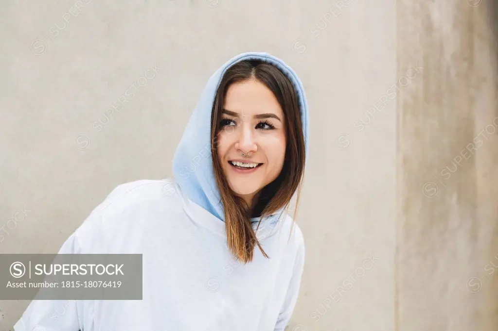 Smiling woman wearing hooded shirt looking away while standing against wall