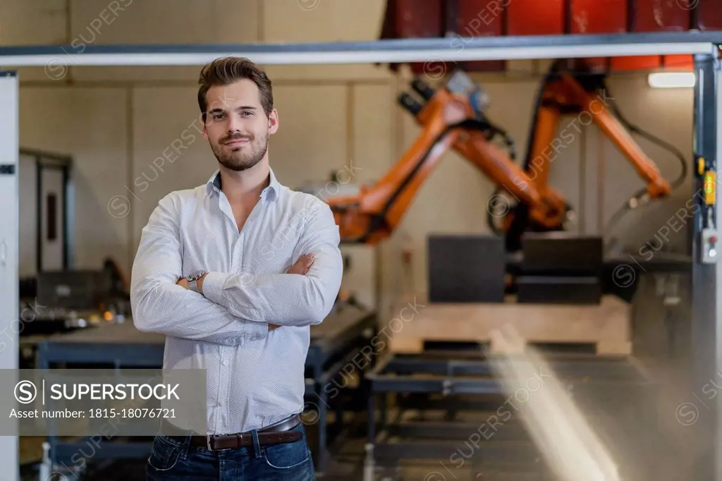 Young businessman standing with arms crossed against robotic arm at factory