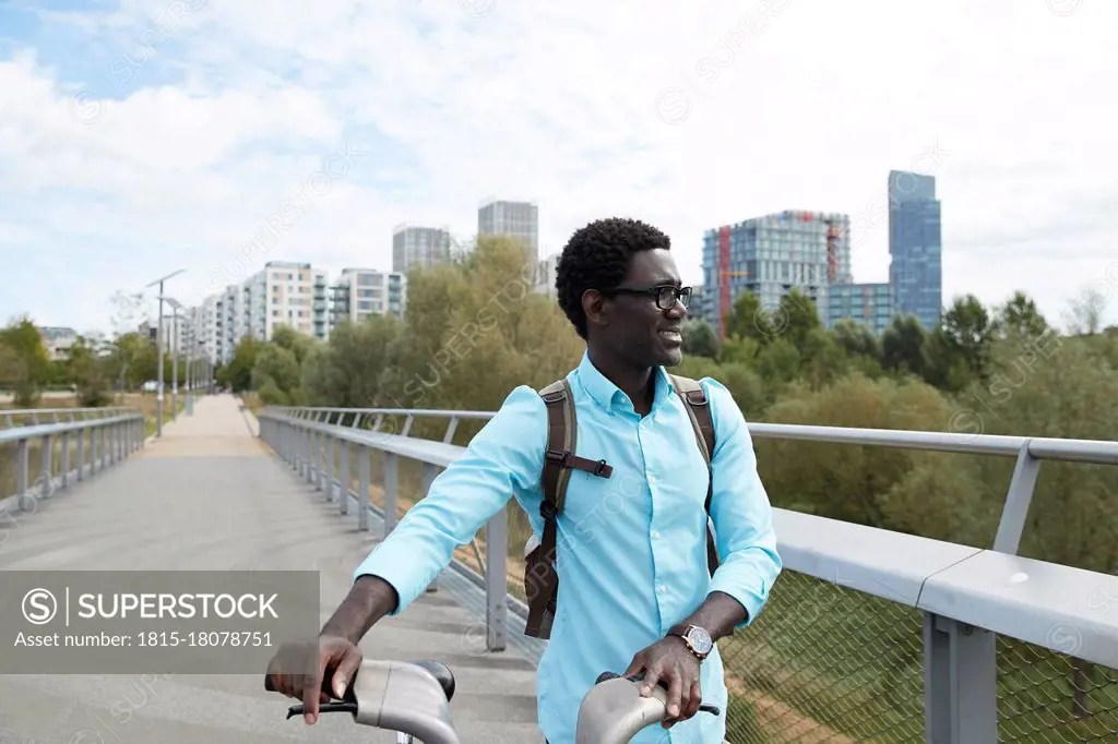 Smiling man looking away while walking with bicycle against sky in city