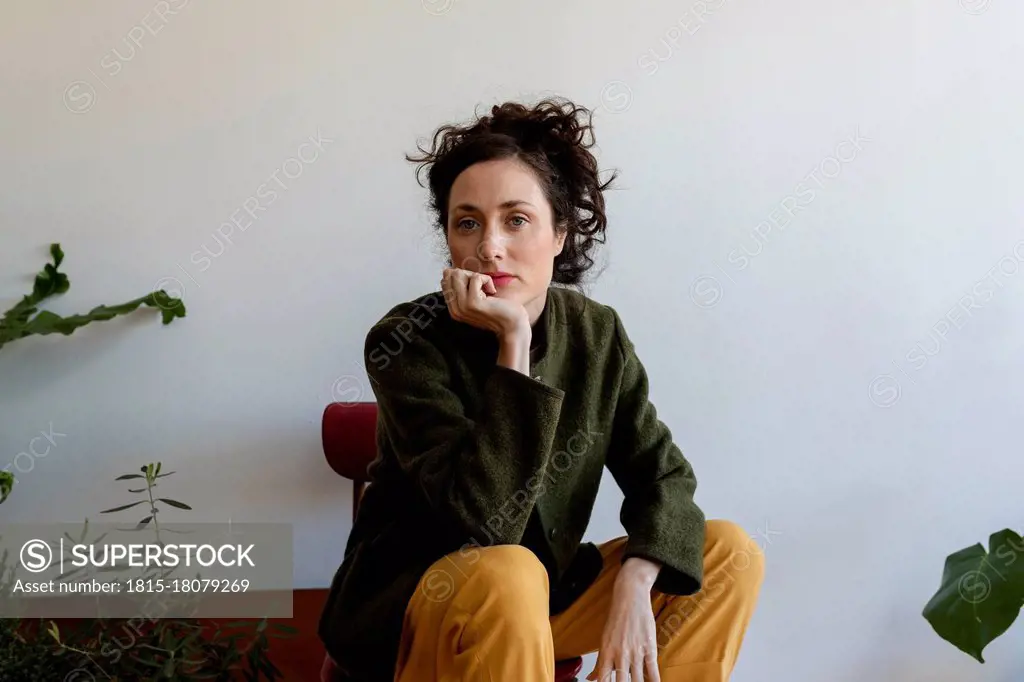 Thoughtful fashionable woman with hand on chin sitting against wall at home