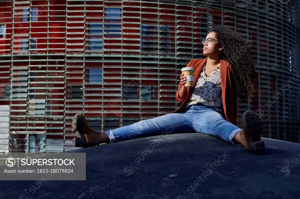 Young woman holding bamboo mug looking away sitting on footpath against building