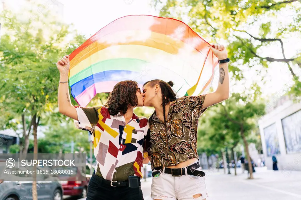 Lesbian couple holding rainbow flag while kissing on footpath in city