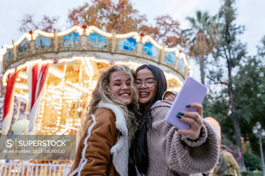 Multi-ethnic female friends talking selfie through mobile phone against illuminated carousel