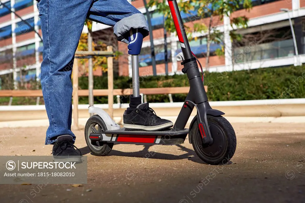 Disabled man with prosthetic leg standing on electric push scooter in playground